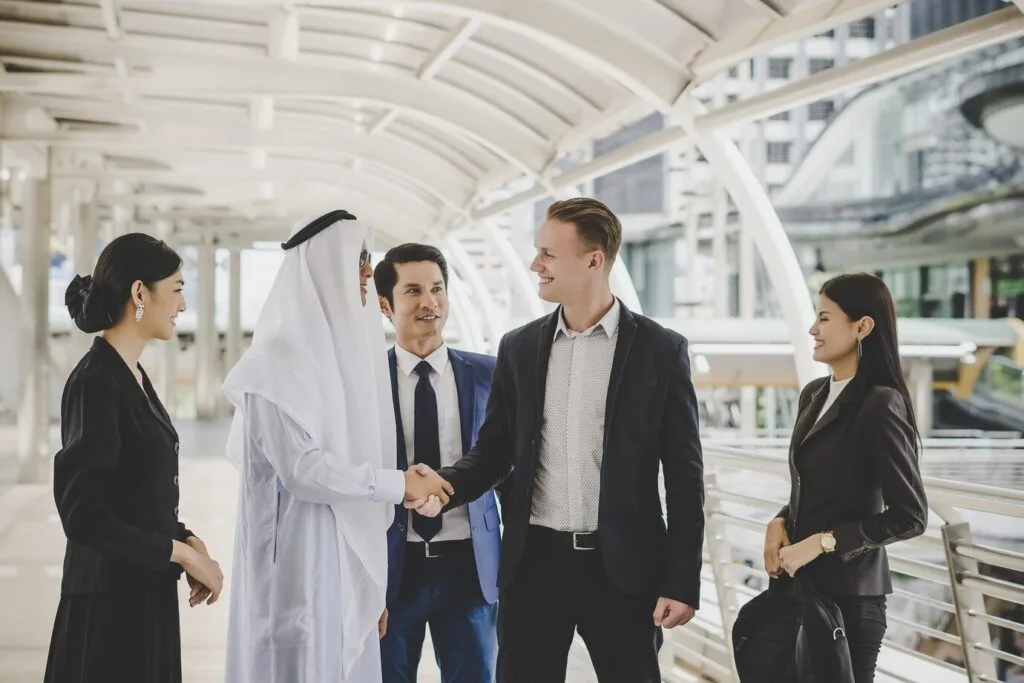 A group of diverse business professionals, including a Saudi individual, engaging in a handshake, representing cross-cultural business collaboration and the importance of effective HR practices in Saudi businesses.